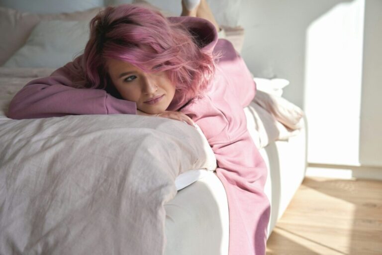 Depressed pensive teen girl introvert pink hair lying on bed looking away.