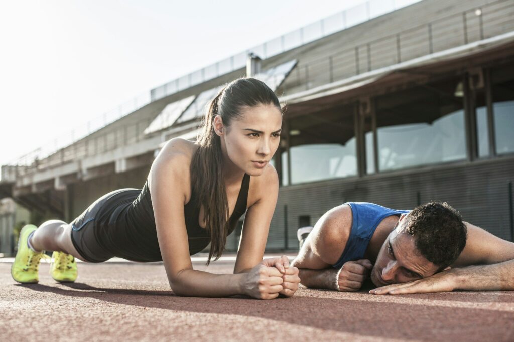 Determined woman doing a plank next to a fatigued man on a track field