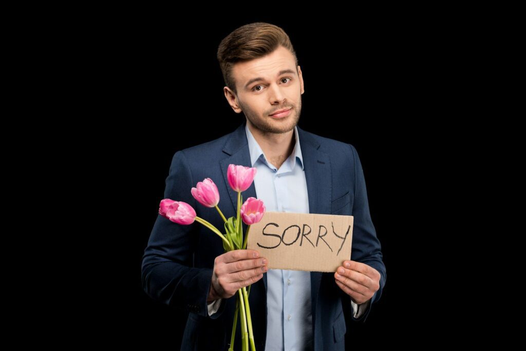 Handsome young man with pink tulips and sorry sign hopefully looking at camera isolated on black