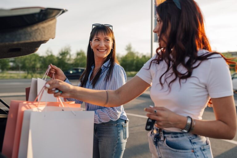 Cheerful trendy female buyers putting shopping bags into a car trunk.